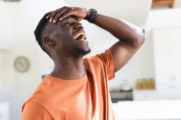 Happy african american man in sunny kitchen, holding head and laughing