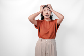 Stressed young Asian woman in a brown shirt and taupe pants is holding her head in her hands...