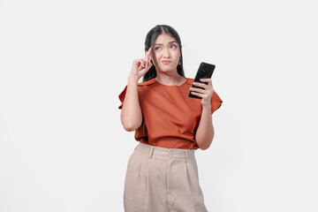 Fototapeta premium Thoughtful young Asian woman in an brown shirt holding a smartphone looking to the copy space aside with a confused expression, isolated by white background.