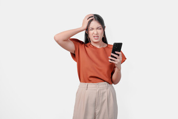 Fototapeta premium Upset young Asian woman holding smartphone and appears to be angry over isolated white background.