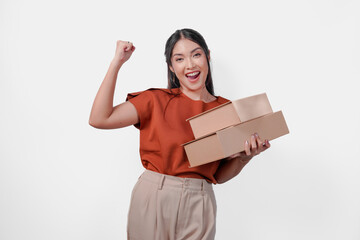 Joyful young Asian woman in a brown shirt is holding gift boxes and raising her arms in the air.
