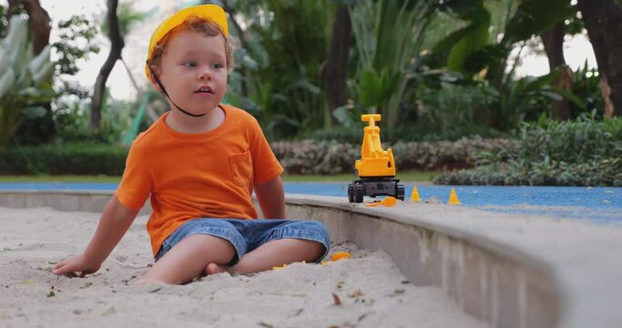 Toddler boy plays as small builder in sandbox, dressed in orange t-shirt and toy safety helmet. Initially upset, he tosses sand aimlessly before calming down and using his hand to scoop and pile sand.