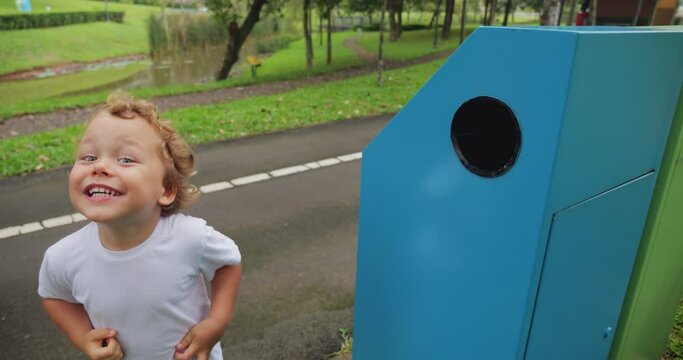 Active toddler boy runs up to dual-bin recycling station and throws two plastic bottles into designated slot. He turns and grins at camera, proudly showing off his accomplishment