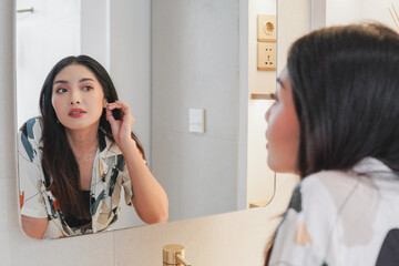 Beautiful young Asian woman wearing patterned shirt looking at herself in the mirror while putting on earring. Beauty concept.