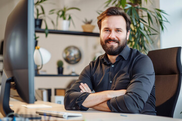 Photo of a modern office showing a bearded businessman sitting at his desk with a computer. He is looking into the camera smiling with his arms crossed in the style of a front view portrait.