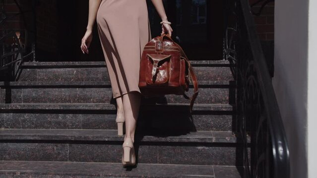 Girl in a dress with a leather travel bag is walking up the stairs, back view