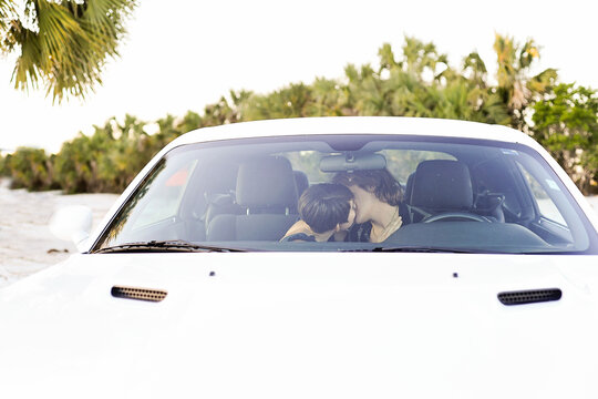 A couple sharing a kiss inside a white car with a tropical backg