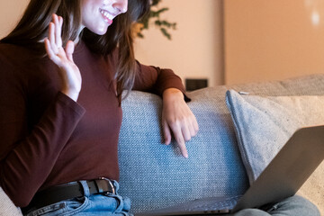Obraz premium Cheerful young adult woman waving hands in front of a laptop by night sitting on sofa. Online dating friends app gadget using computer. One happy female people using notebook late and gesturing hi