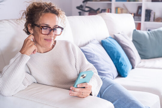 A happy curly lady with glasses relaxes at home alone, sits on the big sofa in a comfortable pose and shares good news on social media via mobile phone. Smiling woman enjoys weekend by ordering food