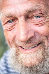Close up portrait of happy 70-year-old optimist man with smiling wrinkled face, a touch of sadness feeling in his blue eyes. Park outdoors background defocused. Elderly people smile at the camera.