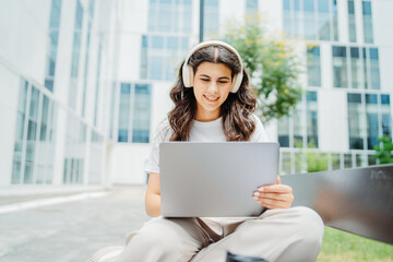 One beautiful young caucasian woman student sitting on bench near university listening to music and studying on laptop	