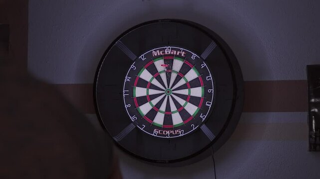 Person throwing darts at a dartboard, hitting triple 20 three times, in a dimly lit room