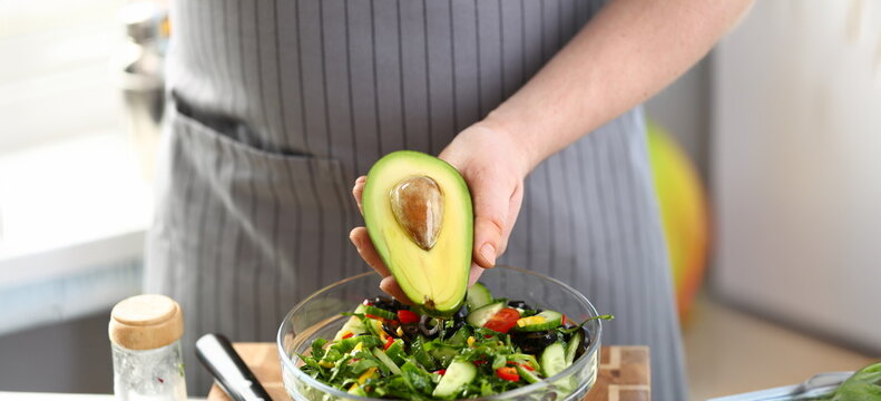 Male Hand Holding Oiled Tropical Avocado Half. Chef Preparing Vegetable Salad with Exotic Fruit Ingredient. Chopped Cucumber, Greens, Tomato and Olives in Glass Bowl Horizontal Photography
