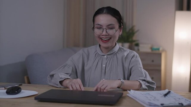 A happy Asian businesswoman sits smiling and gesturing as she has new creative ideas. A laptop and notes are scattered on the table.