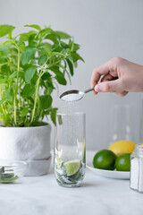 Homemade mojito cocktail drink preparing process, hand holding spoon with sugar, pouring sugar in glass with lime and mint, homemade summer beverage cooking recipe