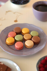 Plate of pastel macarons, cookies and chocolate, cup of tea of coffee, glass of bubble water, various berries, books and accessories on the table. Selective focus, pastel colors.