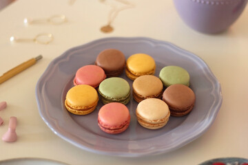 Plate of pastel macarons, cookies and chocolate, cup of tea of coffee, glass of bubble water, various berries, books and accessories on the table. Selective focus, pastel colors.