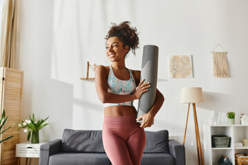 A curly African American woman in active wear holds a yoga mat in a stylish living room, ready for her workout routine.
