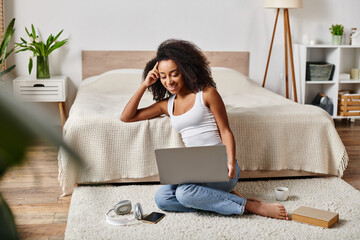 A curly African American woman in a tank top is sitting on the floor, engrossed in using a laptop...