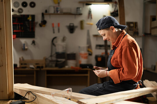 Female Carpenter Resting in Workshop, Sitting on Table Using Smartphone