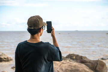 A man wearing a hat is taking photos of the sea view from the beach using a smartphone