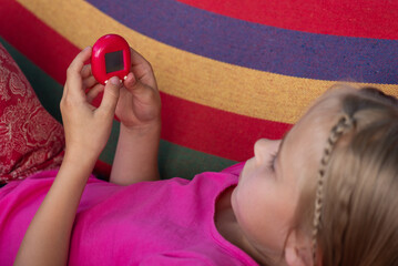 Little girl playing Tamagotchi toy while lying on a striped hammock. Popular game, caring for a...