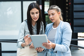 Two coworkers using a digital tablet and talking while standing outside. Modern business colleagues chatting together in front of office building on their way to work. Businesspeople collaboration