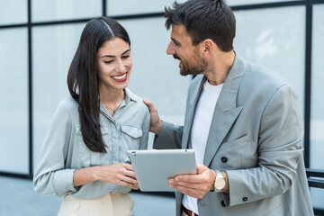 Obraz premium Two coworkers using a digital tablet and talking while standing outside. Modern business colleagues chatting together in front of office building on their way to work. Businesspeople collaboration