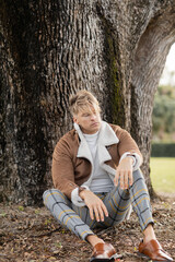 A blonde man in stylish attire leans against a tree in a green Miami park.