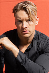 A handsome blonde man in a black shirt poses on the streets with red wall