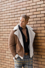 A handsome blonde man in a stylish brown jacket and plaid pants stands against a brick wall