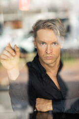 A handsome blonde man in stylish attire, viewed through a window reflection