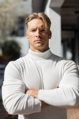 Fototapeta premium A handsome blonde man poses on a Florida street, wearing a white turtleneck sweater and looking directly at the camera.