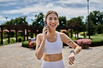 A young woman in activewear, with vitiligo, talks on her phone while smiling in a park.