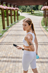 A young woman with vitiligo wears white activewear and a smartwatch as she walks through a park, holding a phone and a water bottle.