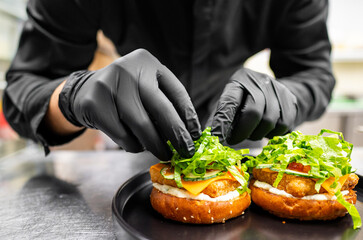 A person in black attire and gloves prepares two sandwiches with lettuce, cheese, and sauce on a black plate