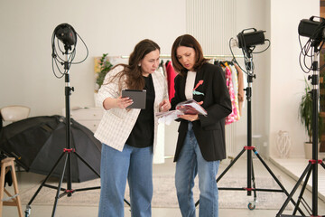 Two bloggers engage in a content discussion, holding a tablet and a fashion magazine while standing in a studio environment with lighting equipment around them. Their focused expressions indicate a