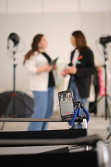 Two fashion bloggers are pictured in a studio, discussing new content ideas with a tablet and a magazine. The presence of lighting equipment suggests a photo or video shoot planning session.