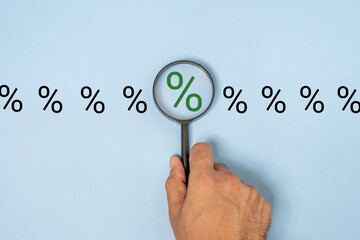Close-up of a hand holding a magnifying glass over a percentage sign on a blue background. Focus, analysis and inspection concept.
