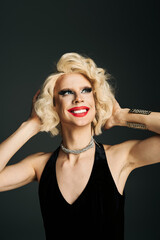 A drag queen with blonde hair and makeup smiles and poses in a black dress against a gray background.