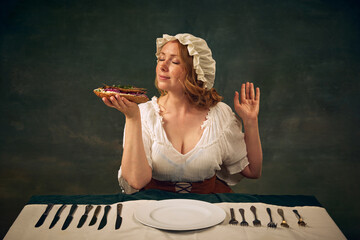 Portrait of young woman medieval clothes sitting at table with plate and enjoying delicious sandwich with fish and onion, set again vintage green background. Concept of food, comparison of eras
