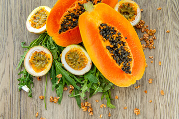 Slices of sweet papaya, maracuja, green rucola surrounded by granola flakes on a wooden background