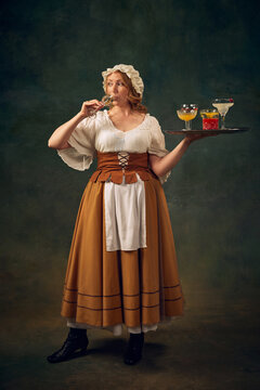 Portrait of woman in historical attire, drinking cocktail and holding a tray with various alcoholic drinks, set against vintage green background. Concept of history, medieval bar, restaurant