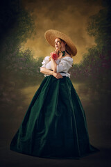 Fototapeta premium Portrait of elegant, beautiful young woman, medieval person in period attire, long skirt, blouse and hat, standing with thoughtful dreamy face against vintage green background. Concept of history