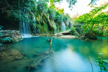 Naklejka premium Kursunlu Waterfall, green lake or pond surrounded by greenery, huge tree roots and stone rocks on a sunny day in Antalya, Turkey.