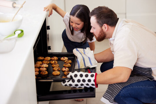 Oven, girl and father with cookies for baking, preparation and checking on tray in home kitchen. Family, dad baker and happy kid for skills development, learning and observation of biscuits by stove