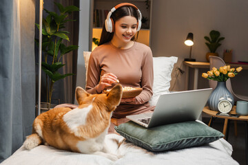 Young girl, teenager wearing headphones watching movie on laptop and eating popcorn with dog on bed