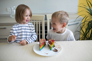 The older brother and little sister are laughing and eating a multicolored cake.
