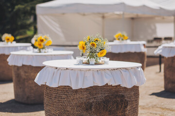 Sunflowers in vase on tables with white tablecloths. Preparation for party in rustic style. A beautiful shot of sunflower decorations. Street cafe table made of straw rolls.