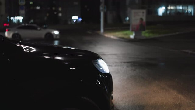 A close-up nighttime view of a black car reversing, with the headlights illuminating the road. Another white car can be seen driving in